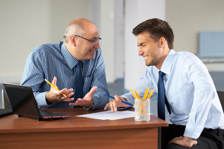 A boss laughs at his computer screen with a male colleague.